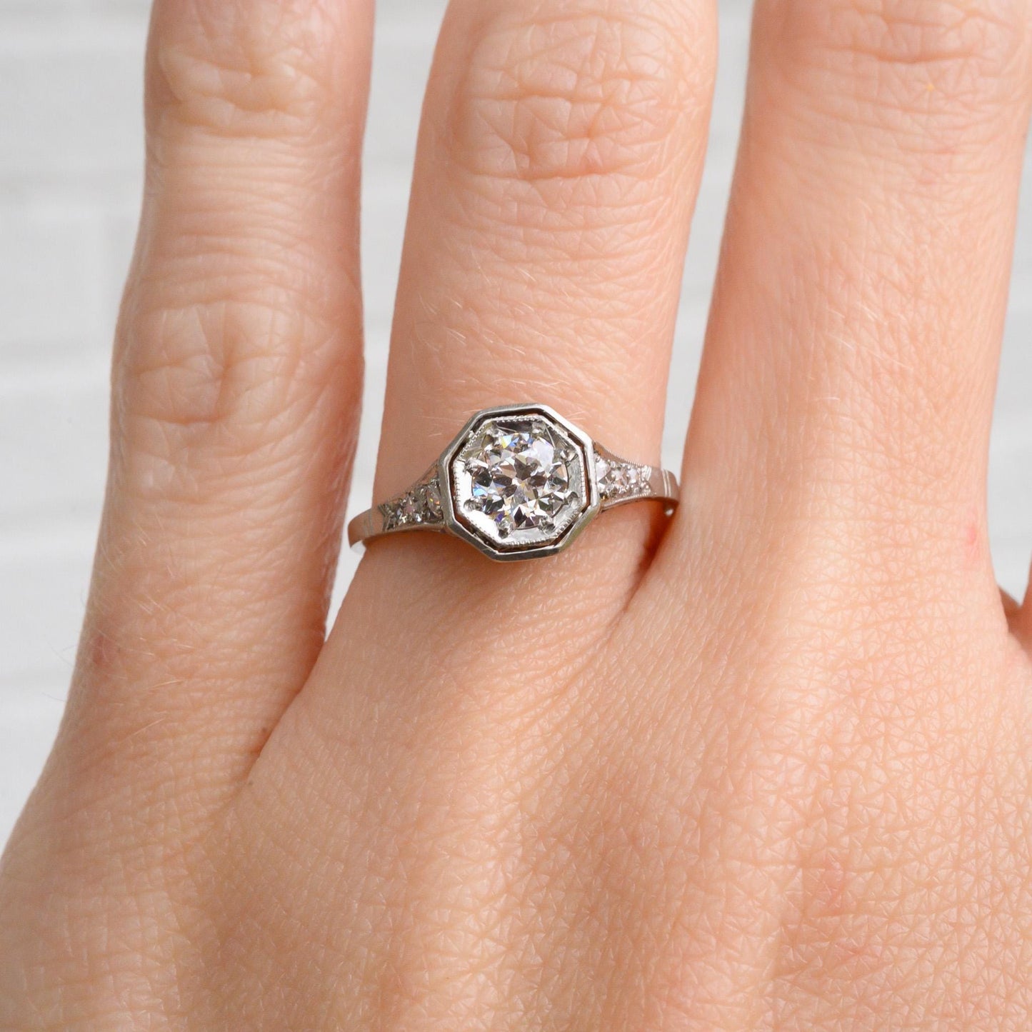 May include: A silver engagement ring with a large octagon-shaped diamond in the center. The diamond is surrounded by smaller diamonds that are set in a pave setting. The ring is on a finger.