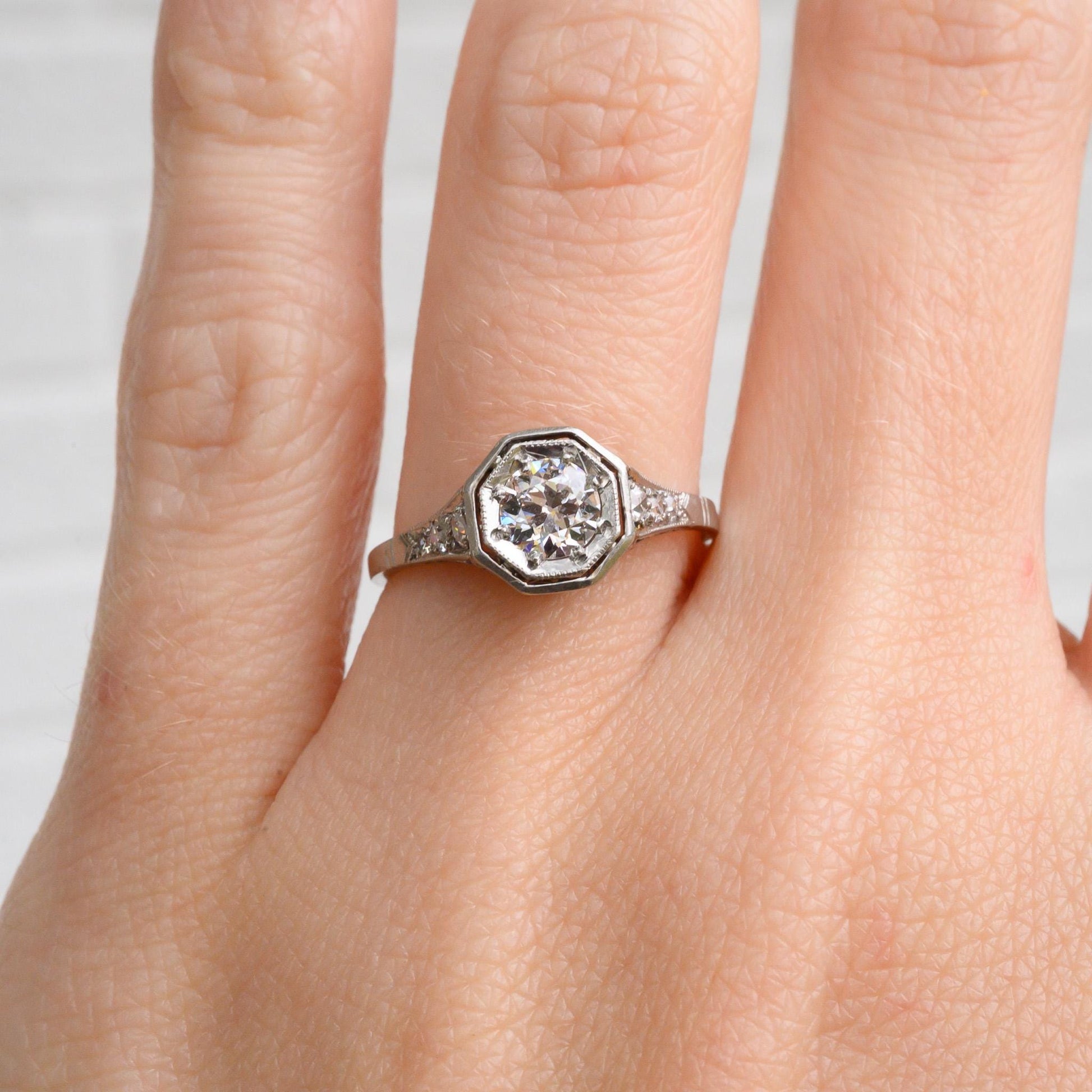 May include: A silver engagement ring with a large octagon-shaped diamond in the center. The diamond is surrounded by smaller diamonds that are set in a pave setting. The ring is on a finger.
