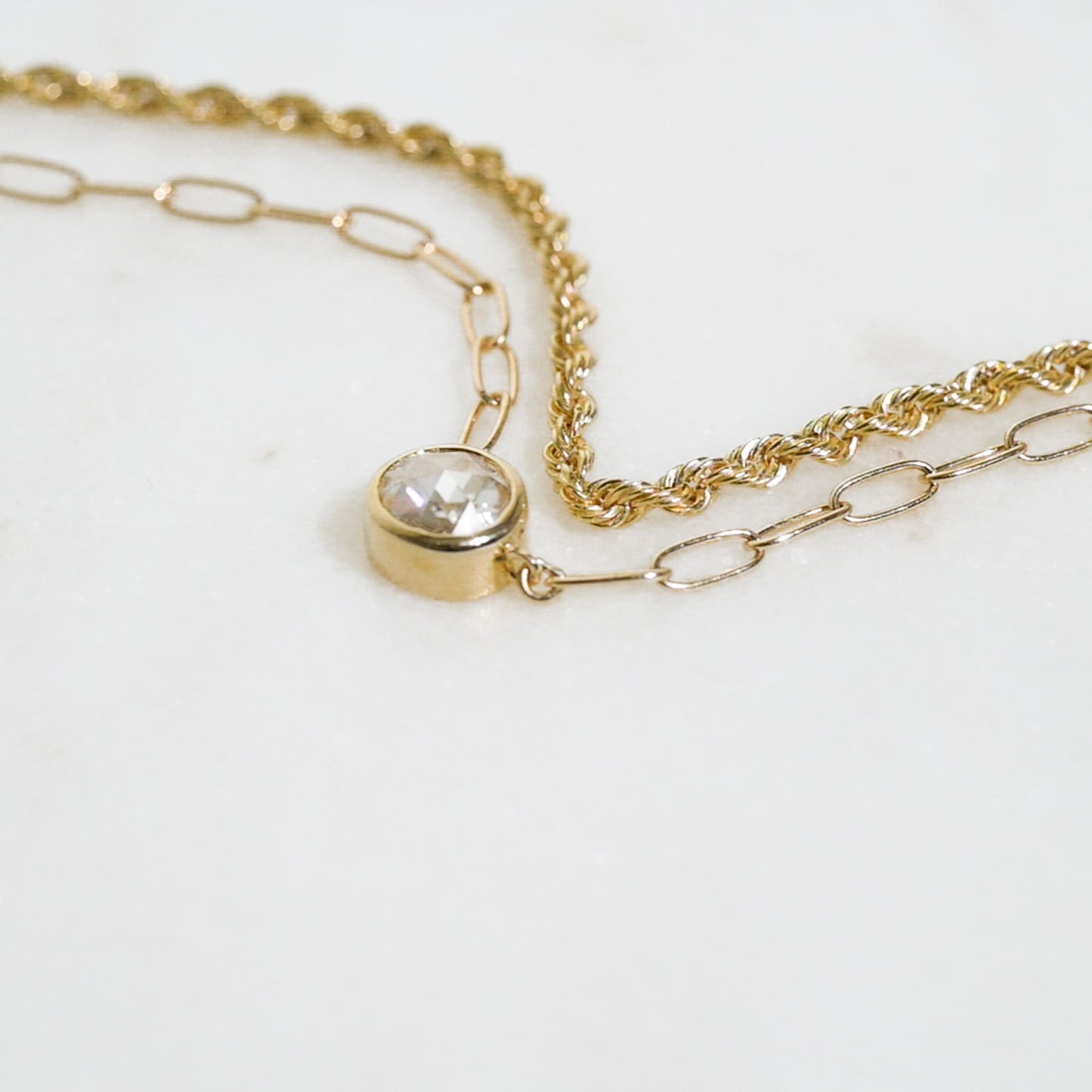 May include: A gold chain necklace with a single round clear gemstone pendant. The chain is made of two different styles, one is a rope chain and the other is a link chain.