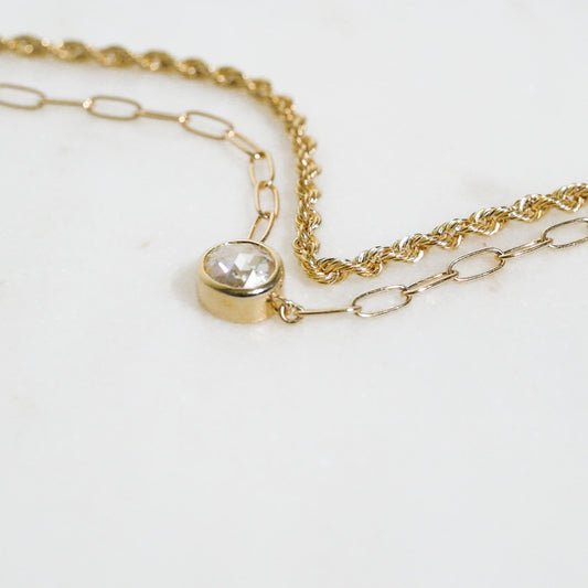 May include: A gold chain necklace with a single round clear gemstone pendant. The chain is made of two different styles, one is a rope chain and the other is a link chain.