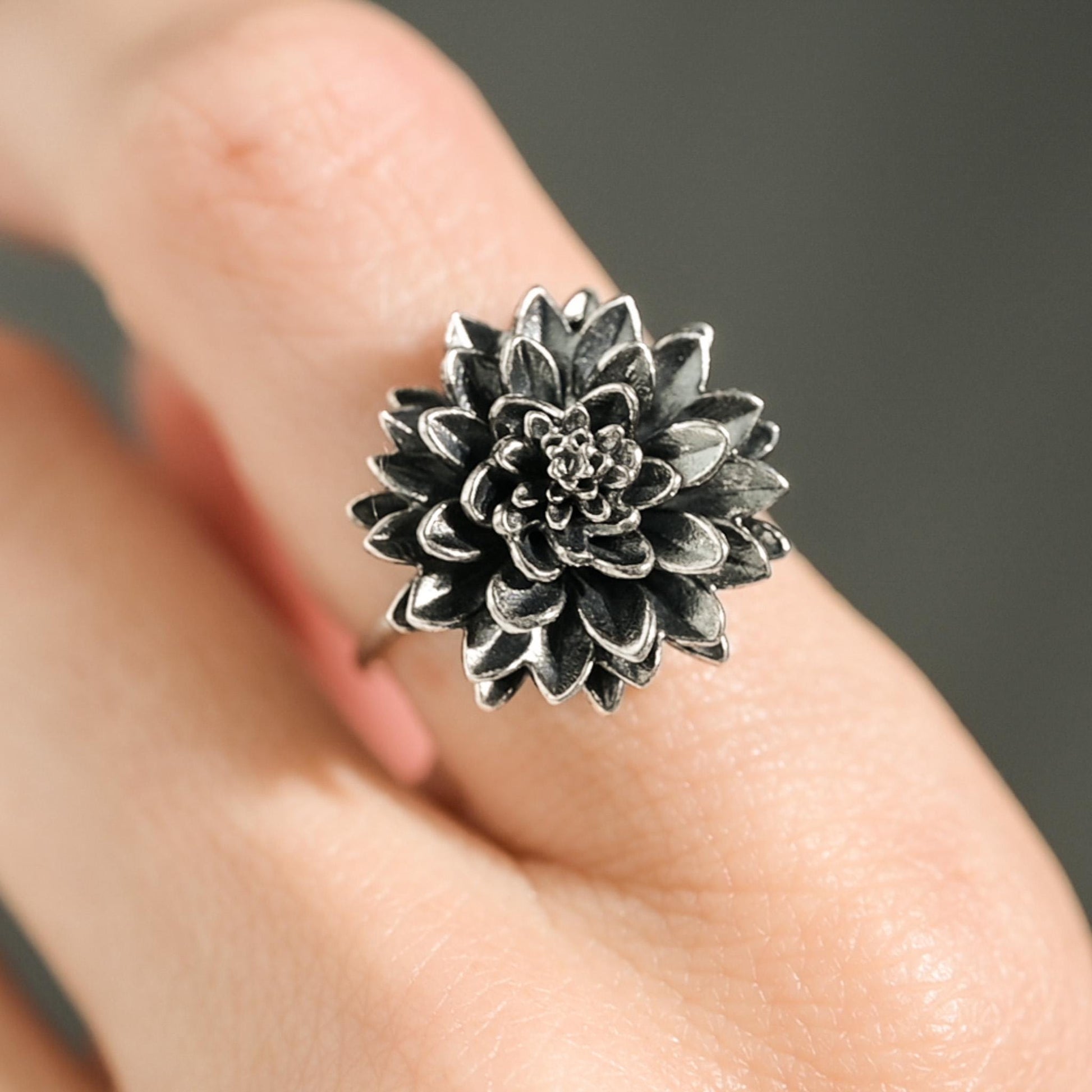 May include: A silver-toned ring featuring a detailed, three-dimensional flower design. The flower has multiple layers of petals, creating a textured appearance. The ring is worn on a finger, showcasing the floral jewelry.